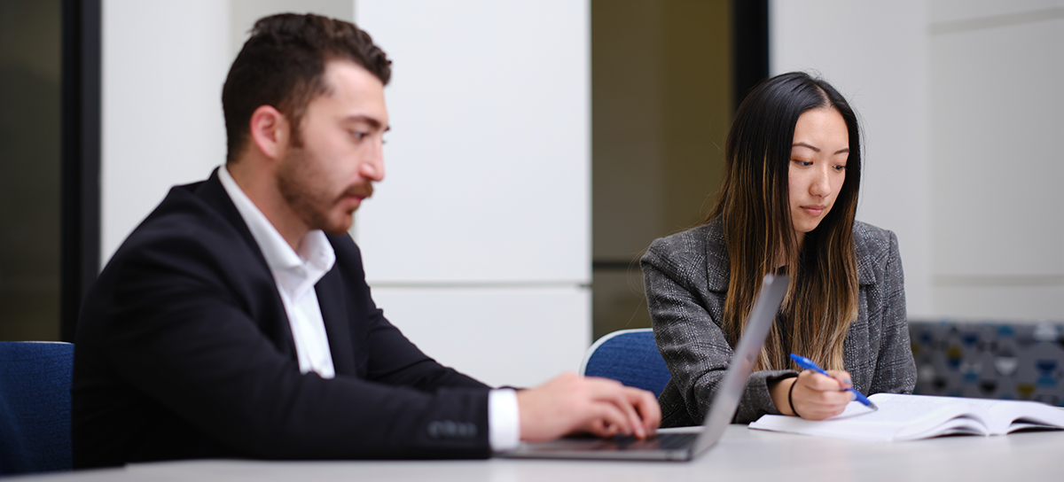 Two students sitting at a table, studying, one at a laptop and one looking at a textbook.